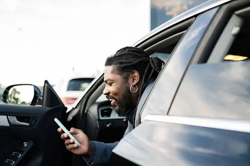 A person with dreadlocks and an earring sits in a car with the door open, smiling while looking at a smartphone—perhaps checking ride details on PerGO NEMT Software.