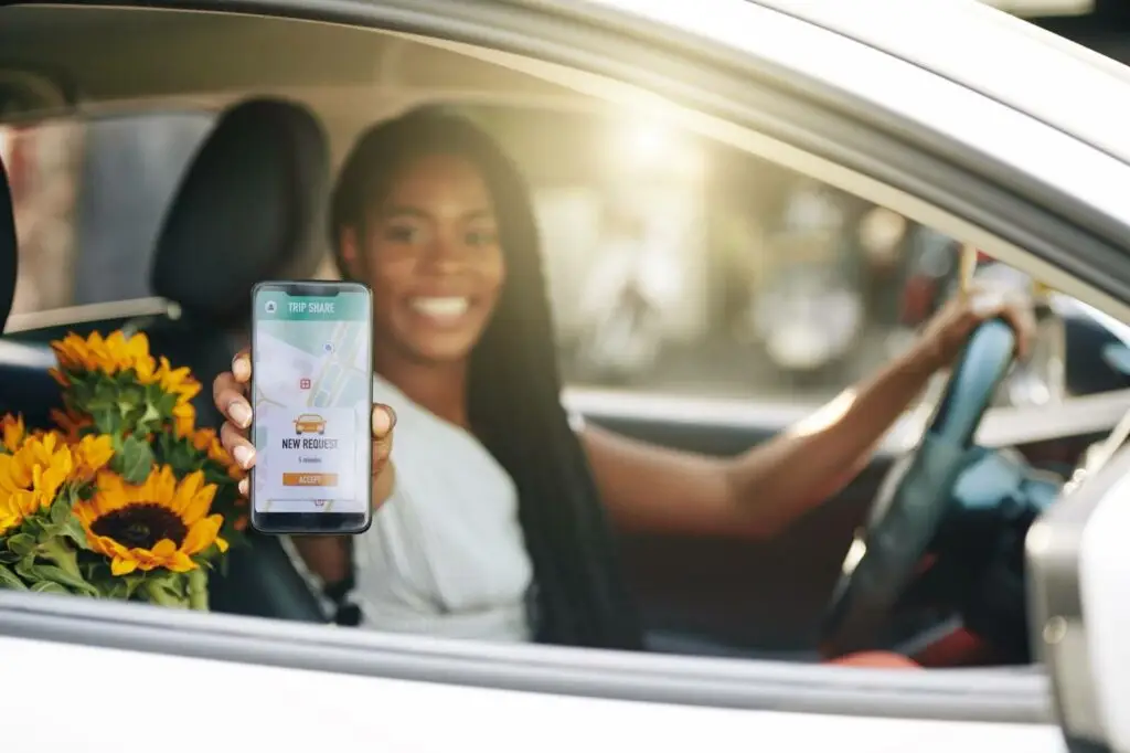 A smiling woman sits in a car’s driver seat, holding a smartphone with a ride-sharing app on the screen. Sunflowers rest on the passenger seat beside her. Sunlight shines through the car window.