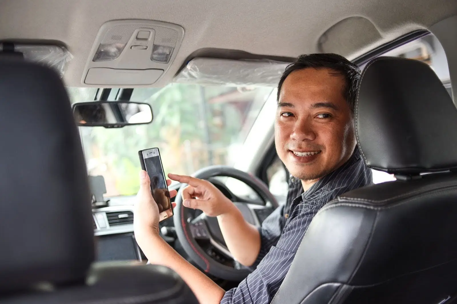 A smiling man sits in the driver&rsquo;s seat of a car, holding a smartphone and pointing at its screen while looking back toward the camera.