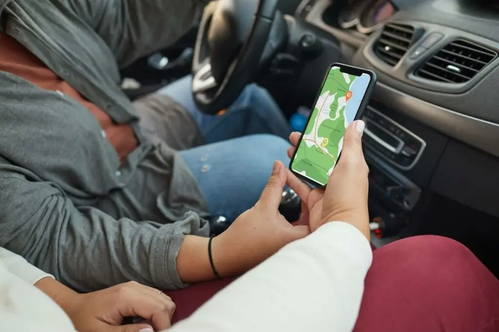 Two people sit in a car, one driving and the other holding a smartphone displaying a map with a location marked, possibly using GPS navigation for directions.