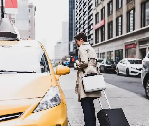 A person in a beige trench coat with a suitcase and bag stands next to a yellow taxi on a city street lined with buildings and parked cars.
