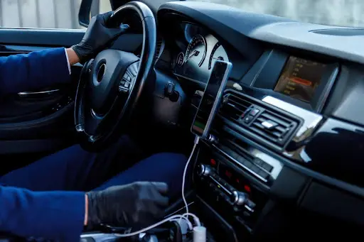 A person wearing black gloves is driving a car. The dashboard displays instruments, a touchscreen, and a smartphone connected with a charging cable—showcasing the modern interior often seen in vehicles using Transportation Management Software.