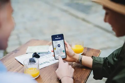Two people sit at a table with drinks and a map, looking at a smartphone displaying a navigation app with a map and location pin. One person is pointing at the phone screen.
