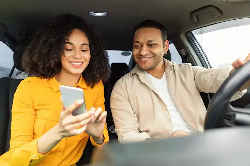 A woman and a man sit in a car; the woman smiles while using a smartphone, and the man, in the driver&rsquo;s seat, looks at her with a smile. Both appear happy and relaxed.