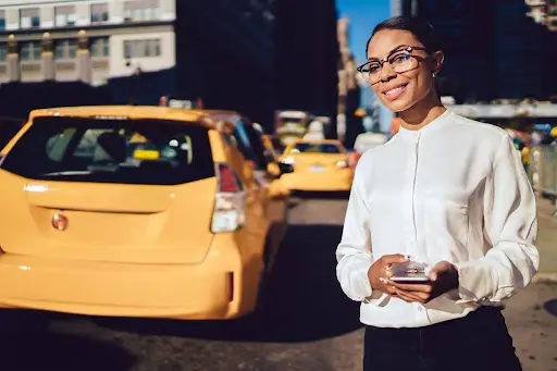 A woman in a white blouse and glasses smiles while holding her phone, standing on a city street with yellow taxis in the background—perhaps using PerGo transportation dispatch software on this sunny day.