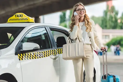 A smiling woman in business attire stands beside a white taxi, talking on her phone and holding a suitcase and a large handbag—perhaps coordinating her ride with PerGo, advanced transportation management software. Trees and a bridge are visible in the background.