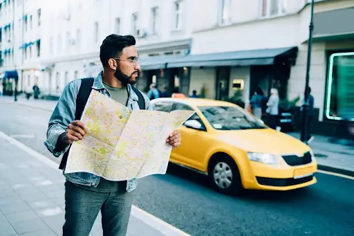 A man with glasses and a backpack stands on a city sidewalk, holding an open map and looking to the side, while a yellow taxi—possibly managed by transportation dispatch software—passes by on the street behind him.