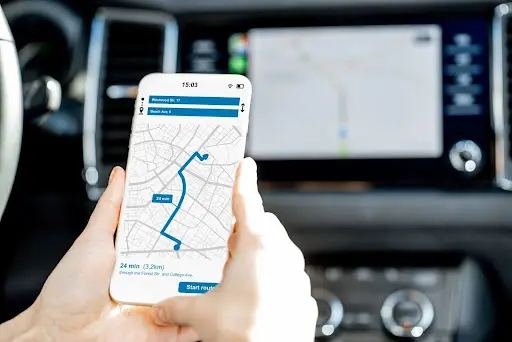 A person holds a smartphone showing a GPS navigation route inside a car, with the car's dashboard and built-in navigation screen—featuring PerGo Transportation Management Software—visible in the background.