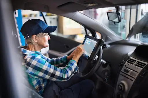 An older person wearing a blue cap and plaid shirt sits in a car, using a diagnostic tablet connected to the vehicle&rsquo;s dashboard&mdash;perhaps maintaining it for use with transportation dispatch software.