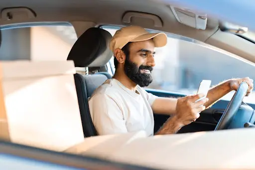A smiling man with a beard and cap sits in a car, holding a smartphone in one hand and the steering wheel in the other, using Transportation Management Software, with cardboard boxes visible on the passenger seat.