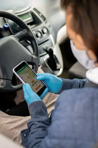 A person wearing blue gloves and a face mask sits in a car, holding a smartphone displaying NEMT Software with ride-hailing features, including a map and booking options on the screen.