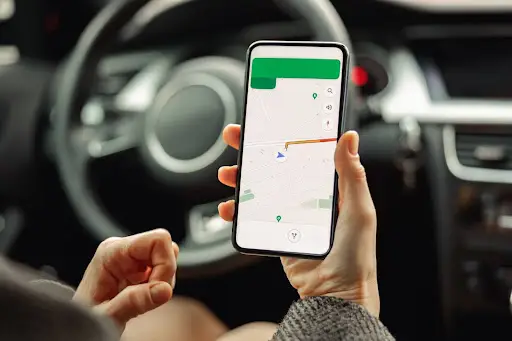 A person sitting in a car holds a smartphone displaying the PerGo transportation management software, with a route highlighted on the screen and the car&rsquo;s steering wheel visible in the background.