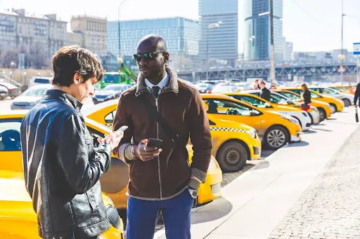 Two men stand on a sidewalk near a row of yellow taxis. One checks his phone, while the other, wearing sunglasses, looks ahead holding his phone&mdash;efficiently managing rides with PerGo&rsquo;s Transportation Management Software amid the bustling cityscape.