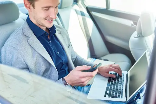 A man sits in the back seat of a car, smiling while using PerGo transportation management software on his smartphone and laptop. He is wearing a blue shirt and a light gray blazer.
