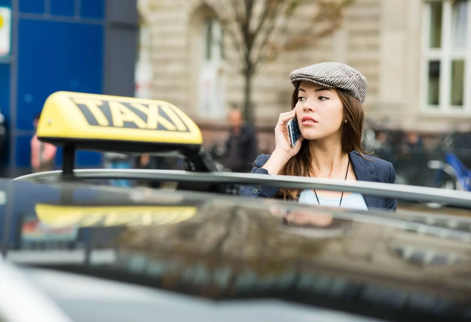 A young woman in a flat cap stands by a taxi, talking on her phone, with a city street and blurred buildings in the background&mdash;perhaps coordinating her ride with transportation dispatch software.