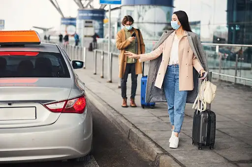 A woman wearing a face mask hails a taxi at an airport curb while pulling a suitcase, highlighting the need for efficient NEMT Software. Another masked person with luggage stands behind her, looking at a phone.