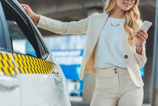 A woman in a light-colored suit stands next to a white taxi, holding a smartphone in one hand and resting the other on the car roof&mdash;efficient travel made easy with PerGo transportation dispatch software. The background is blurred.
