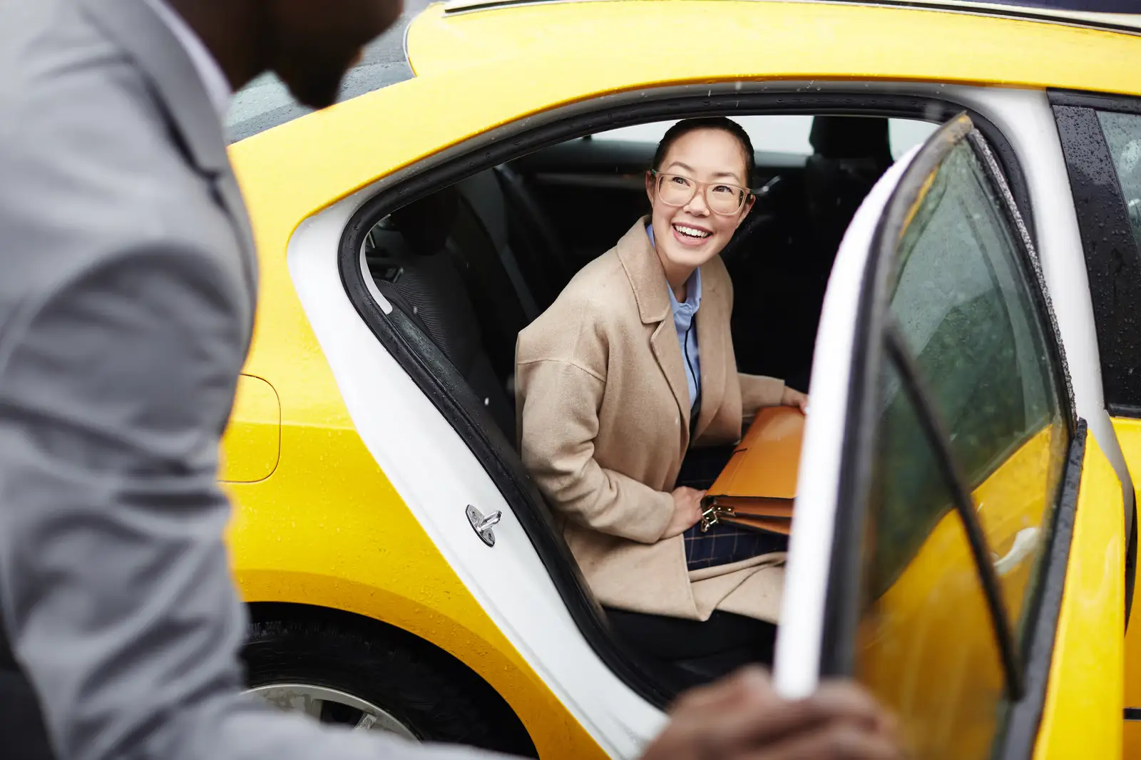 A woman wearing glasses and a beige coat smiles while sitting in the backseat of a yellow car, holding an orange folder&mdash;showcasing how NEMT Software like PerGo makes trips smoother. A person in a gray jacket stands by the open car door.