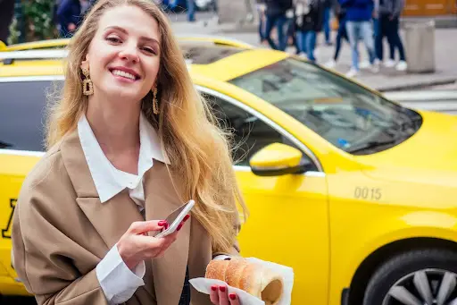 A smiling woman with long blonde hair stands in front of a yellow taxi, holding a phone—perhaps using transportation dispatch software—in one hand and a pastry in the other. She is wearing a tan coat over a white shirt, with people and city buildings visible in the background.
