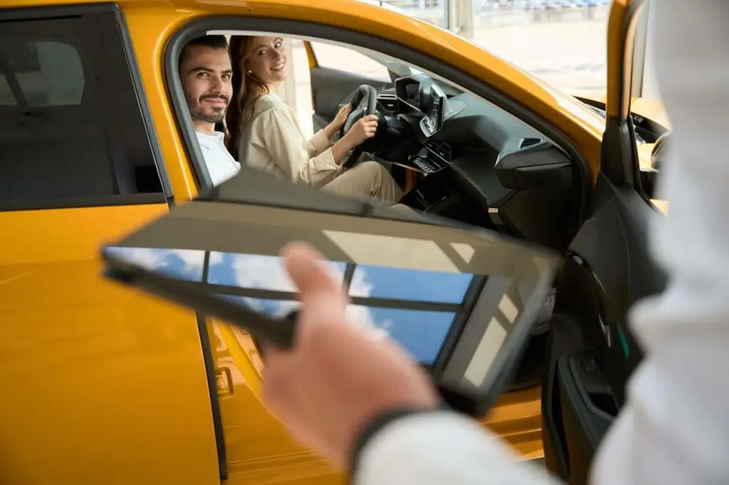 A man and woman sit in the front seats of a yellow car, smiling, while a person holding a tablet—possibly using PerGo transportation dispatch software—stands outside the open driver’s door, likely at a car dealership.
