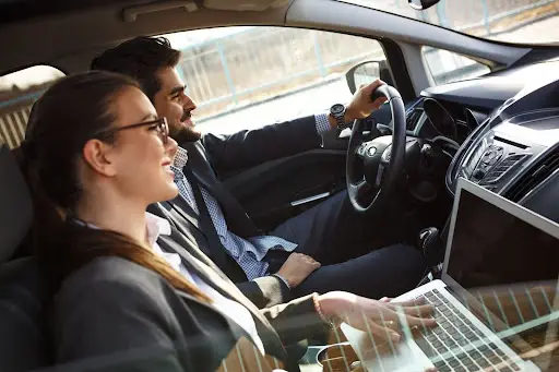 Two business professionals in a car; one is driving while the other works on a laptop featuring transportation dispatch software. Both are dressed in business attire and appear engaged and focused.