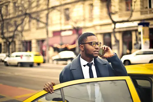 A man in a suit and glasses stands by an open yellow car door, talking on his phone—perhaps coordinating with PerGo transportation dispatch software—with a city street and blurred buildings in the background.