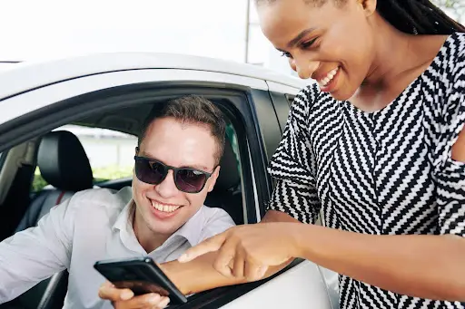 A smiling man wearing sunglasses sits in a car while a woman standing outside shows him something on her smartphone—both appear happy and engaged, possibly exploring transportation dispatch software together.