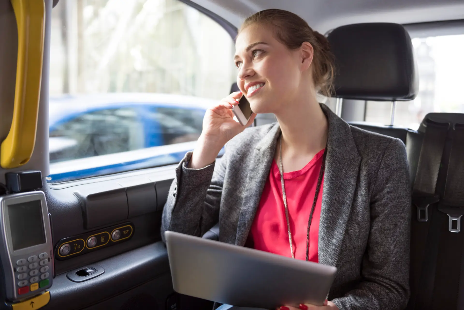 A woman in a gray blazer and red blouse sits in the back seat of a car, smiling while talking on her phone and holding a laptop on her lap. The background shows a blurred view outside the car window.