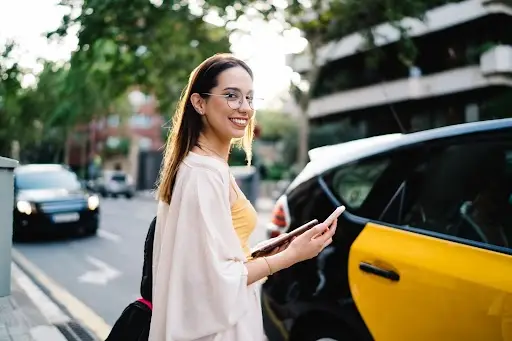 A young woman with glasses smiles while standing next to a black and yellow taxi on a city street, holding her phone and a tablet, with cars and trees in the background.